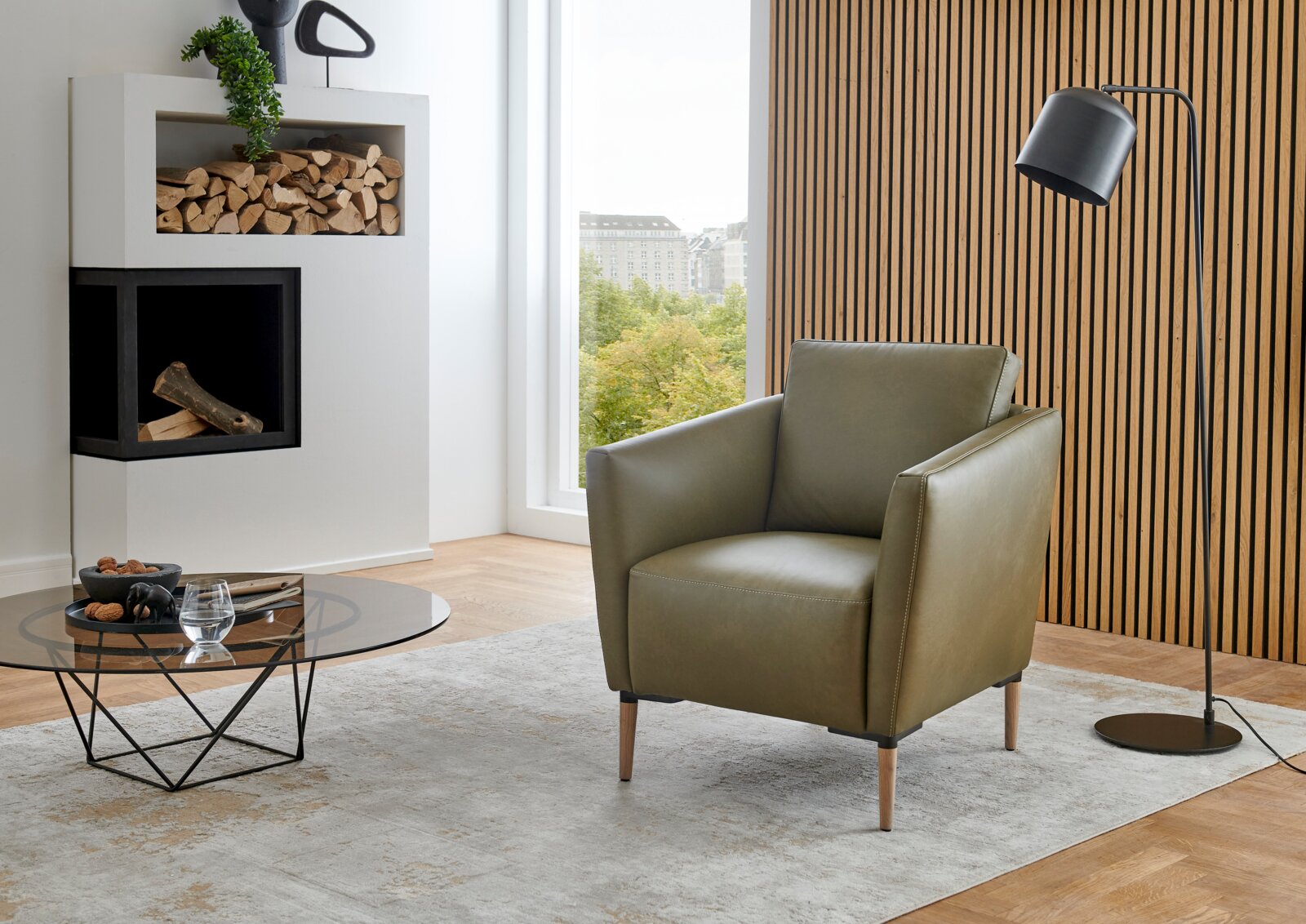 Moderner grüner Sessel Valnatura Polstermöbel PERLA in einem stilvollen Wohnzimmer mit Holzvertäfelung und Stehlampe, seitliche Perspektive.