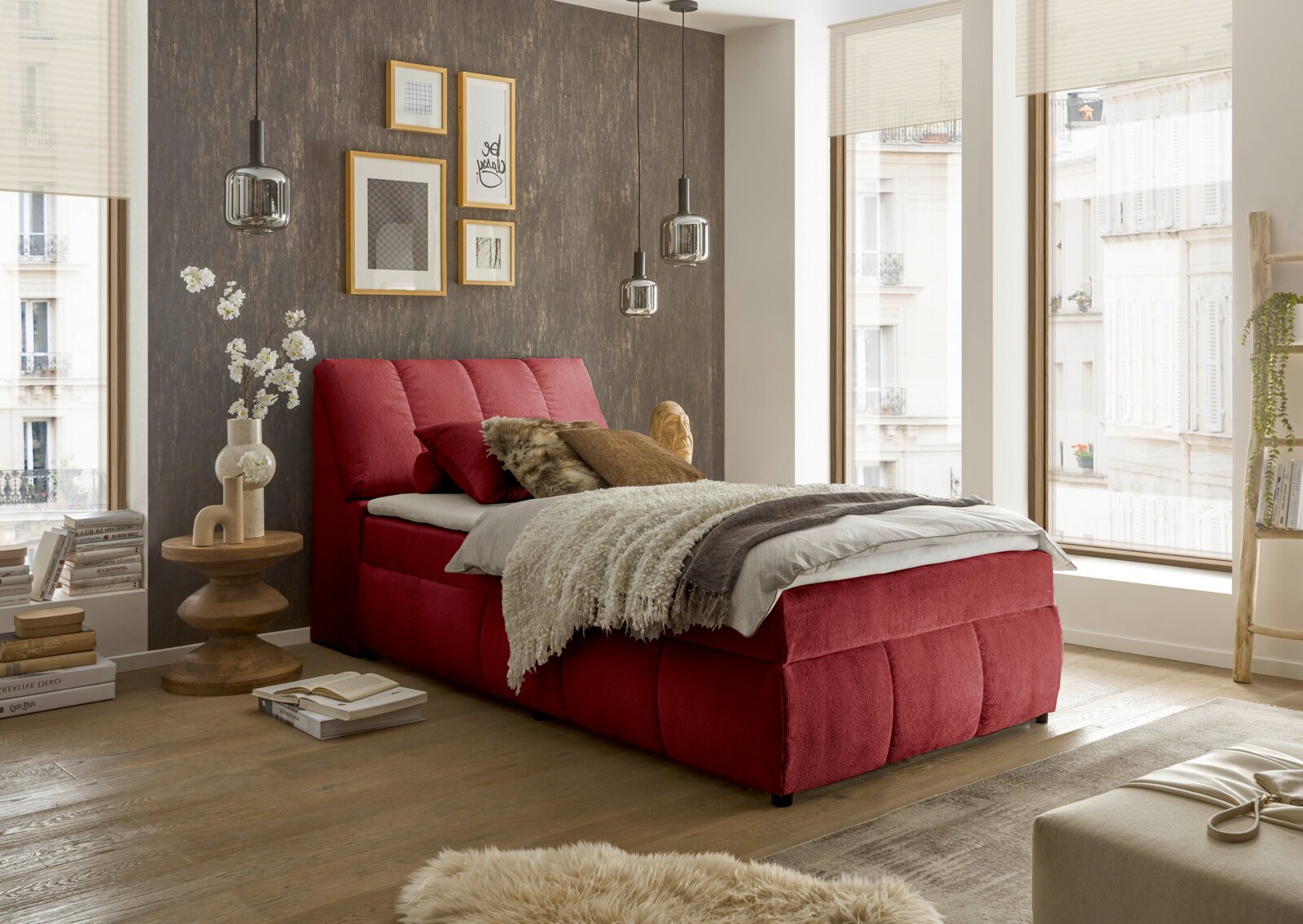Rood boxspringbed in een stijlvol ingerichte slaapkamer met houten vloer en grote ramen, zijaanzicht