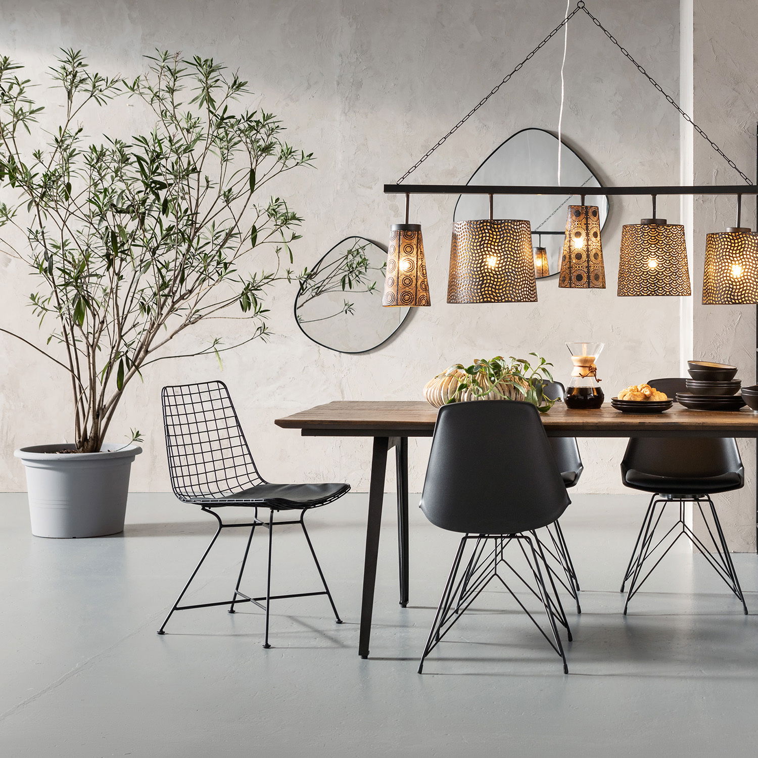 Moderner Essbereich mit einem schwarzen Drahtstuhl im Vordergrund, seitliche Perspektive. Der Raum ist stilvoll mit einem Holztisch, schwarzen Stühlen und hängenden Lampen dekoriert.