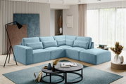 Modernes hellblaues L-förmiges Ecksofa mit breiter Ottomane und vielen Rückenkissen in einem hellen Wohnzimmer, aufgenommen aus frontaler leicht seitlicher Perspektive.