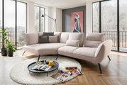 Moderne Longchairkombination in einem hellen Wohnzimmer mit großen Fenstern, die eine Aussicht auf die Natur bieten. Die Perspektive zeigt das Sofa von vorne, umgeben von einem runden Teppich und einem Couchtisch mit Zeitschriften und einem Krug Orangensa