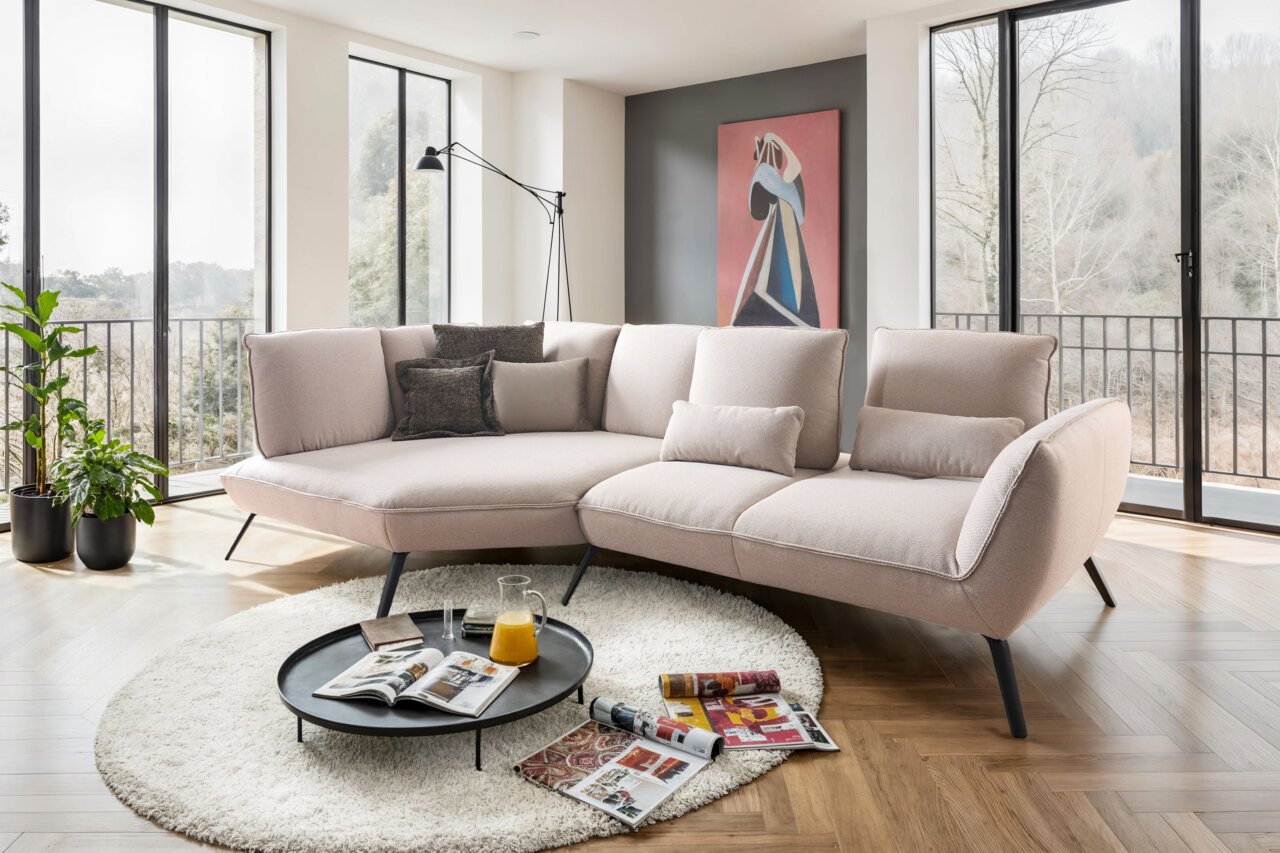 Moderne Longchairkombination in einem hellen Wohnzimmer mit großen Fenstern, die eine Aussicht auf die Natur bieten. Die Perspektive zeigt das Sofa von vorne, umgeben von einem runden Teppich und einem Couchtisch mit Zeitschriften und einem Krug Orangensa