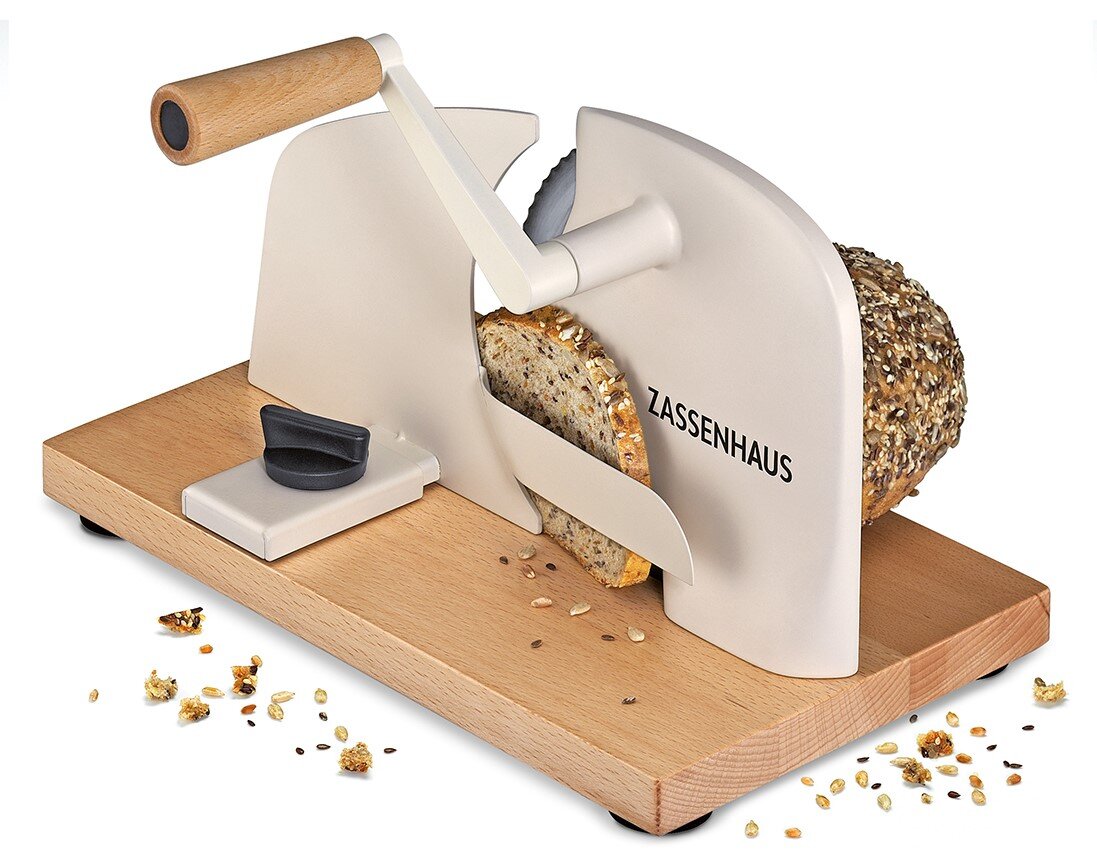 Zassenhaus handmatige broodsnijmachine met wit metalen frame en houten basis, snijdt een meergranenbrood schuin vooraanzicht op een witte achtergrond.