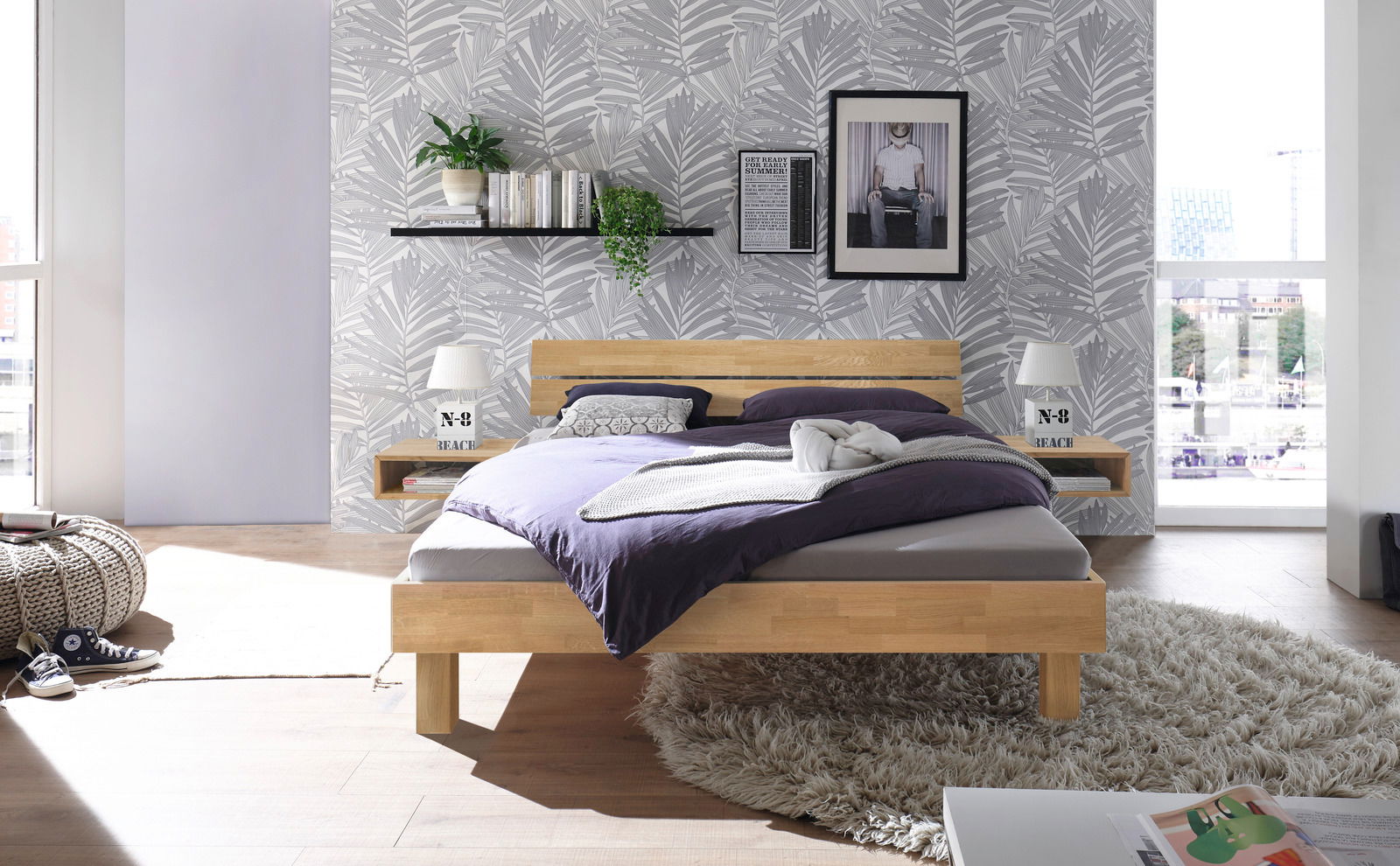 Modernes Schlafzimmer mit einem Kopfteil aus Holz und Metall, frontal fotografiert. Das Bett ist mit lila Bettwäsche bezogen und steht auf einem flauschigen Teppich. Die Wand ist mit einem Blattmuster tapeziert und dekoriert mit Pflanzen und Bildern.