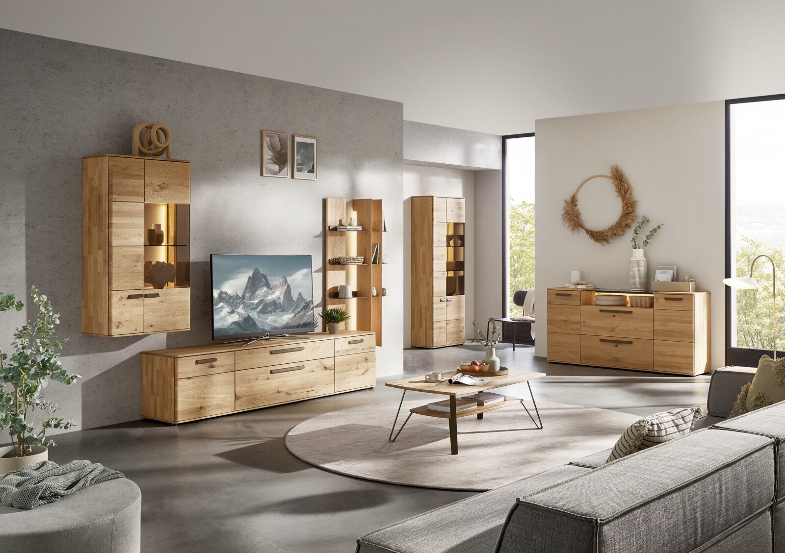 Valnatura Wandkombination LERIDA Modernes Wohnzimmer mit Wohnwand und Sideboard aus hellem Holz mit schwarzen Griffen, teils mit beleuchteter Vitrine, TV-Board und offenen Regalen; aufgenommen in schräger Frontansicht aus leicht erhöhter Perspektive.