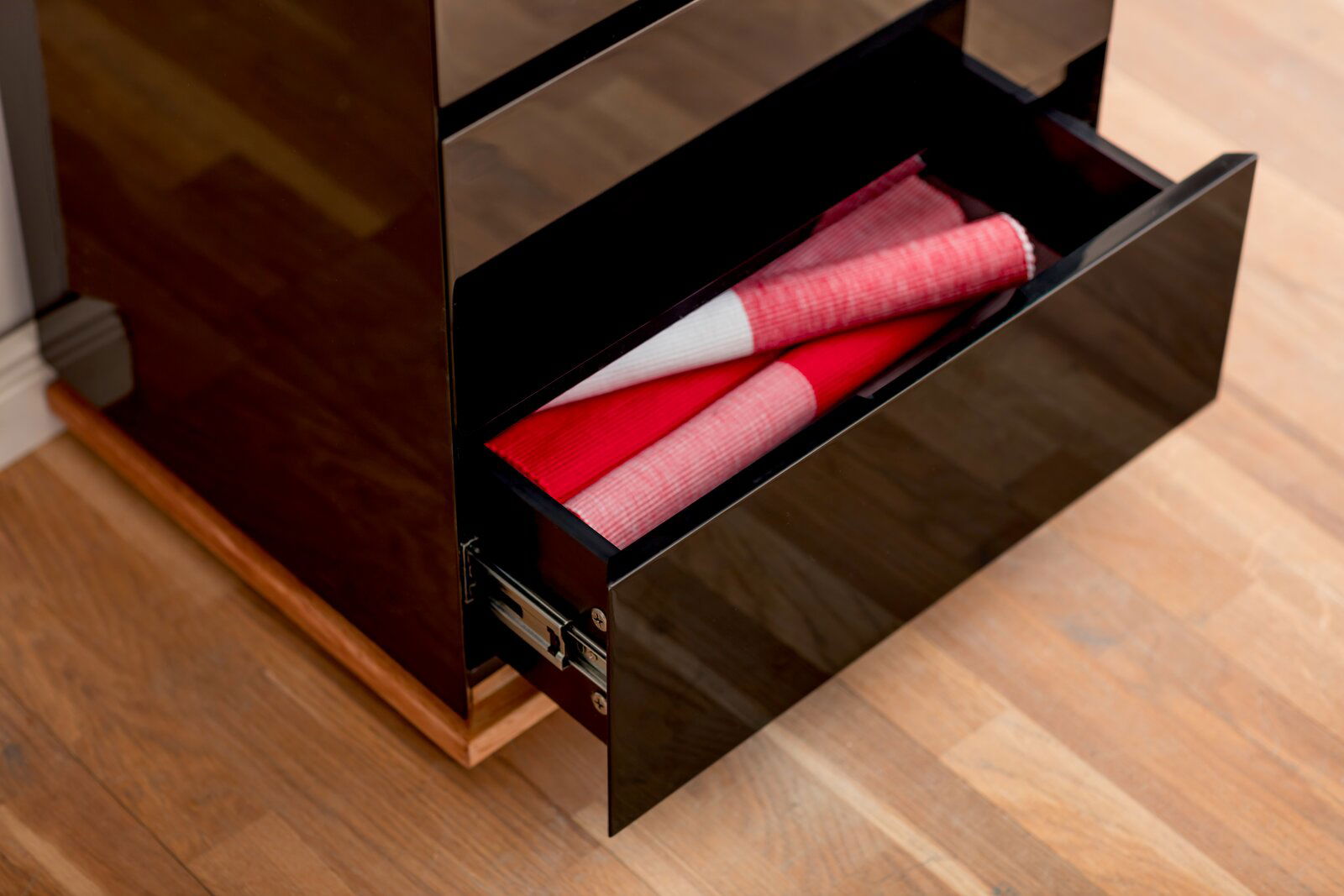 Nahaufnahme eines schwarzen Hochglanz-Sideboards mit geöffneter Schublade, die rote und weiße Stoffe enthält, auf einem Holzboden.