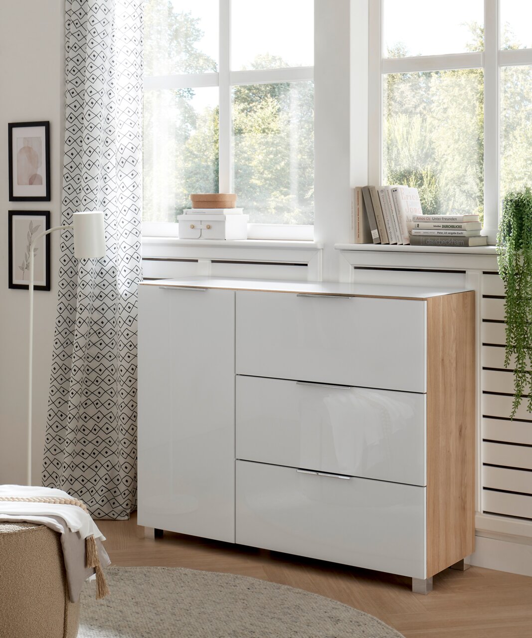 Moderne Kommode mit weißer Hochglanzfront und Holzseiten, in einem hellen Raum mit großen Fenstern, aus seitlicher Perspektive fotografiert.