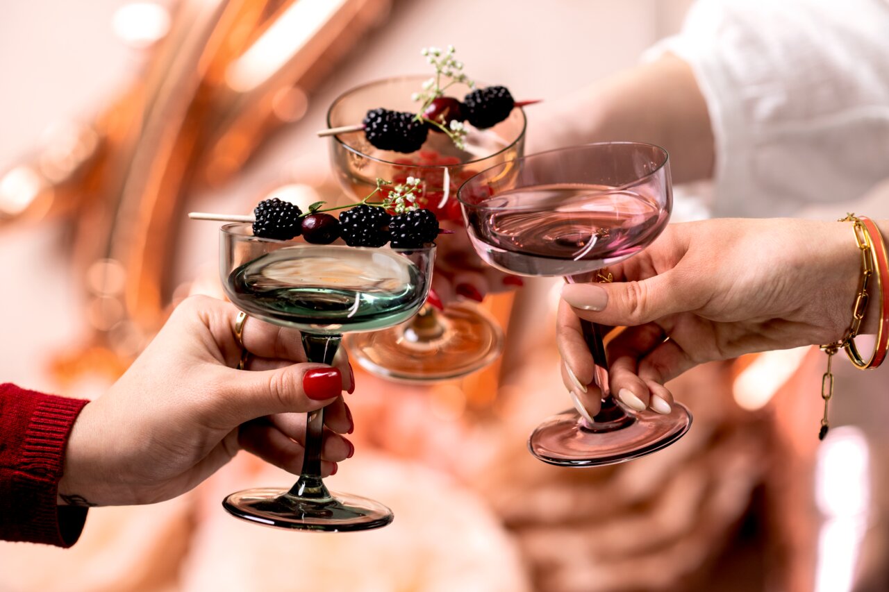Drei Sekt- und Dessertschalen in verschiedenen Farben, gefüllt mit Beeren und dekoriert mit Blumen, werden von Händen in einer fröhlichen Atmosphäre gehalten. Perspektive: Nahaufnahme von schräg oben.