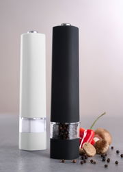 Zwei Gewürzmühlen in modernem Design, eine schwarz und eine weiß, gefüllt mit Pfefferkörnern und Salz, auf einer grauen Oberfläche mit frischen Gewürzen im Vordergrund. Seitliche Perspektive.