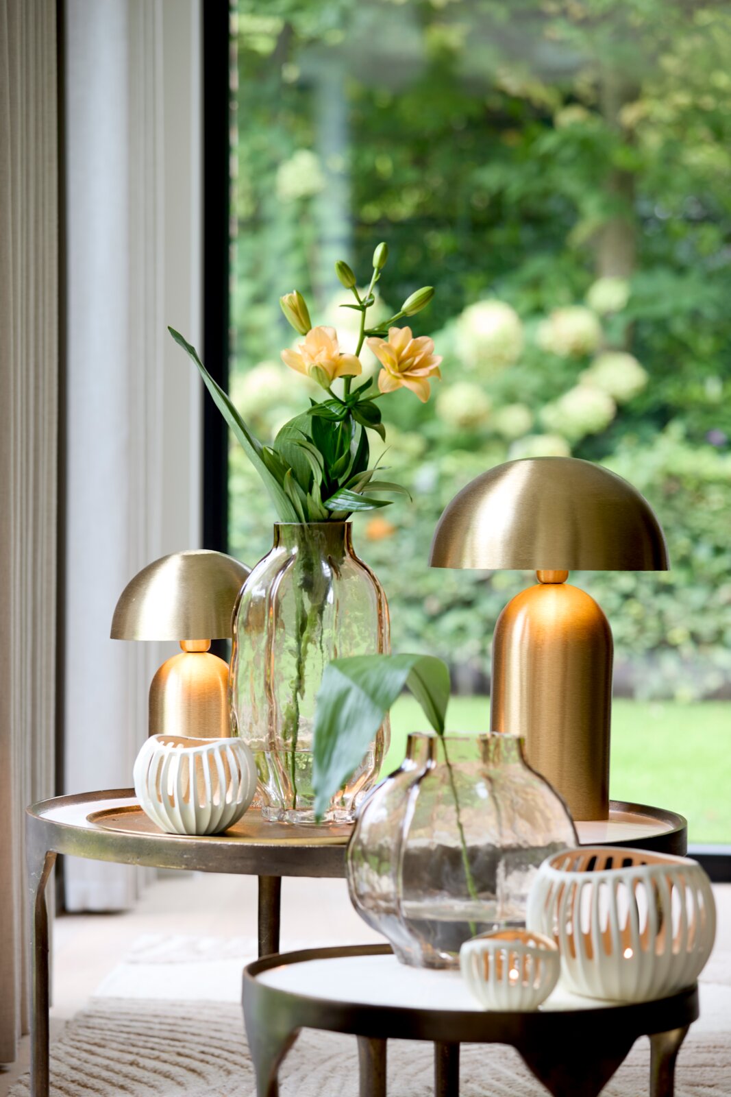 Sfeervolle goudkleurige tafellampen, glazen vazen met bloemen en witte windlichten op ronde bijzettafels, gefotografeerd vanuit vooraanzicht in een licht interieur met tuinzicht.