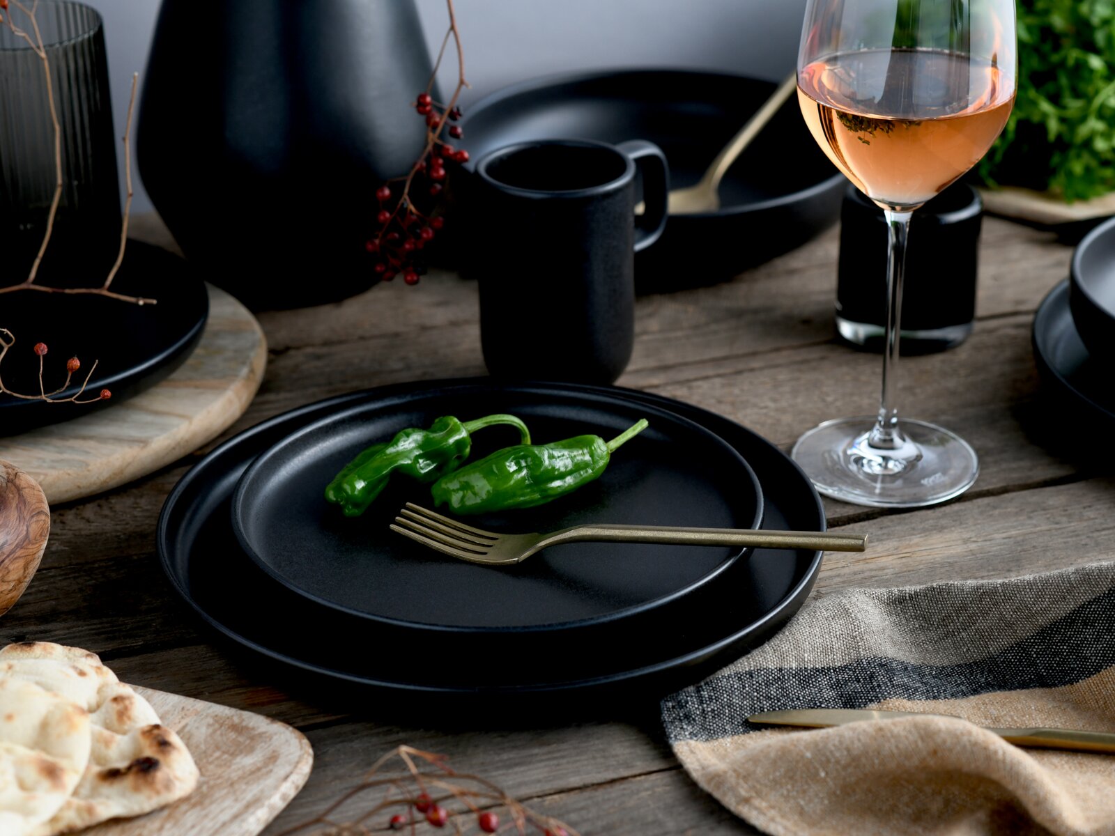 Schwarzes, mattes Geschirr-Set mit Tellerstapel und Becher auf rustikalem Holztisch, dazu goldene Gabel und grüner Paprika sowie ein Glas Roséwein; aufgenommen aus leicht erhöhter, schräger Vorderansicht.