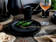 Schwarzes, mattes Geschirr-Set mit Tellerstapel und Becher auf rustikalem Holztisch, dazu goldene Gabel und grüner Paprika sowie ein Glas Roséwein; aufgenommen aus leicht erhöhter, schräger Vorderansicht.