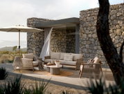 Moderne Loungegarnitur-Set auf einer Terrasse mit Steinmauer im Hintergrund, bestehend aus einem Sofa, zwei Sesseln, einem runden Tisch und einem Sonnenschirm, seitliche Perspektive.