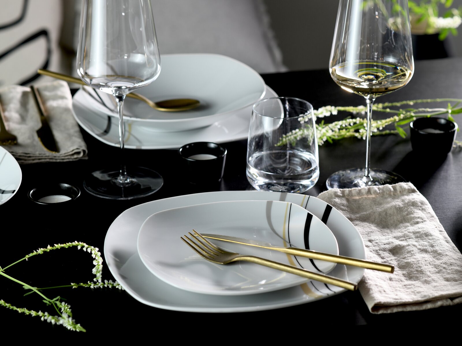 Modern gedeckter Esstisch mit weißem Geschirr mit schwarzen und goldenen Linien, goldfarbenem Besteck, Wein- und Wassergläsern sowie Leinenservietten auf schwarzer Tischplatte, perspektivisch in leichter Draufsicht.