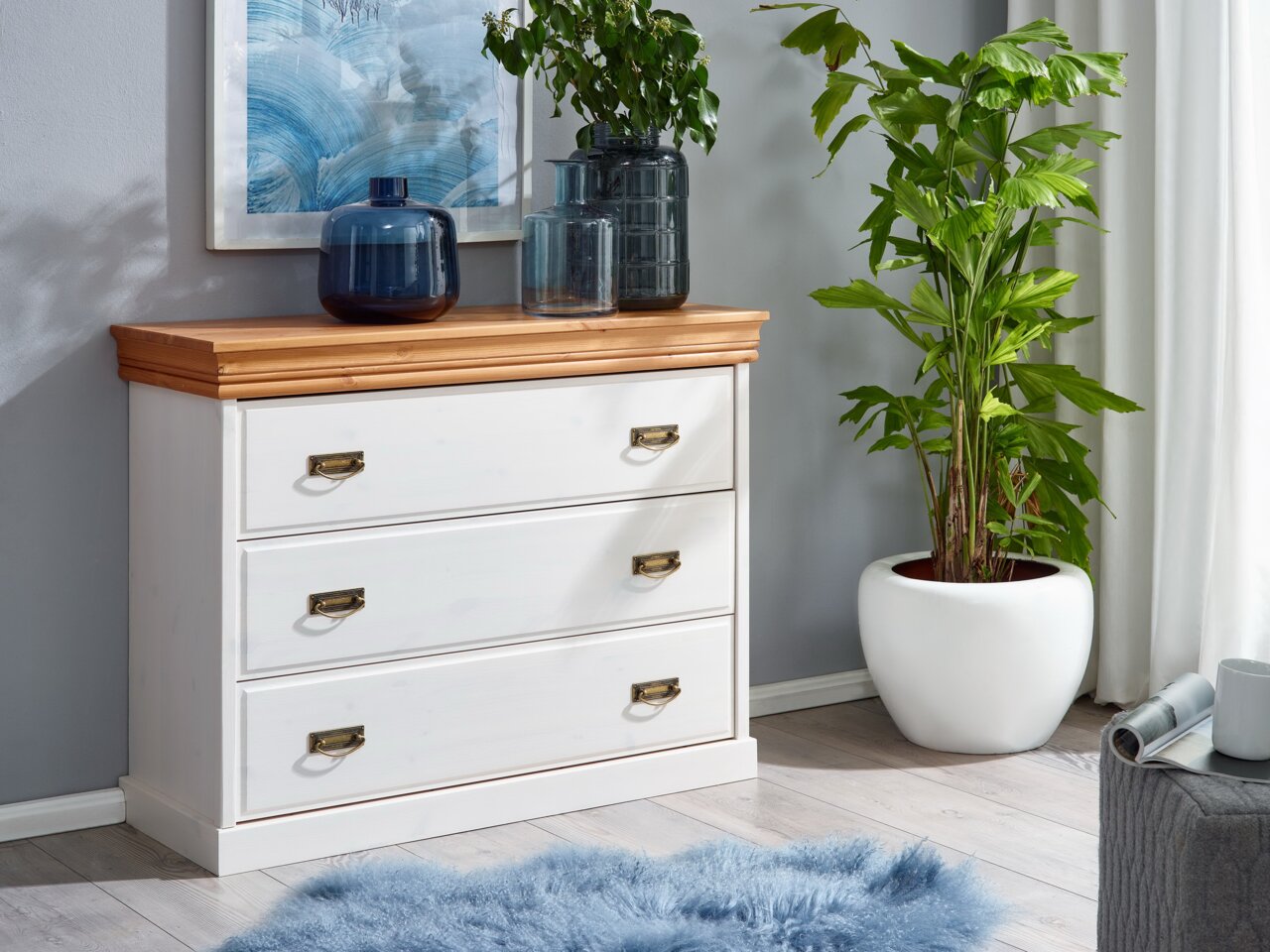 Weiße Kommode mit drei Schubladen und Holzplatte, dekoriert mit blauen Glasvasen, in einem modernen Wohnzimmer mit Pflanzen und Kunstwerk an der Wand, seitliche Perspektive.