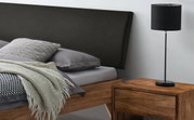 Zijaanzicht van een modern bedframe met een donker hoofdeinde, grijs beddengoed en een houten nachtkastje met een zwarte lamp.