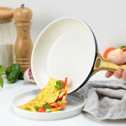 Bratpfanne mit goldfarbenem Griff, aus der ein Omelett auf einen Teller gleitet, seitliche Perspektive.