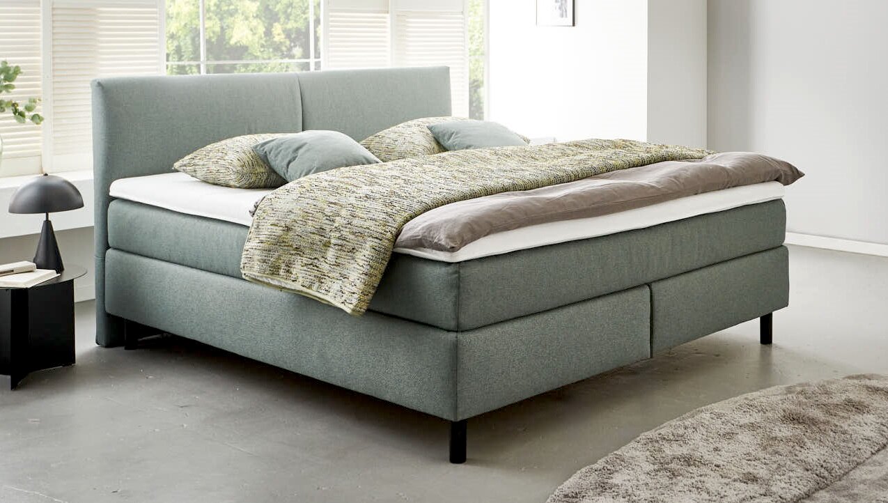 Boxspringbett BX 180/200 in modernem Design, seitliche Perspektive, mit grauem Stoffbezug und gemütlicher Bettwäsche.