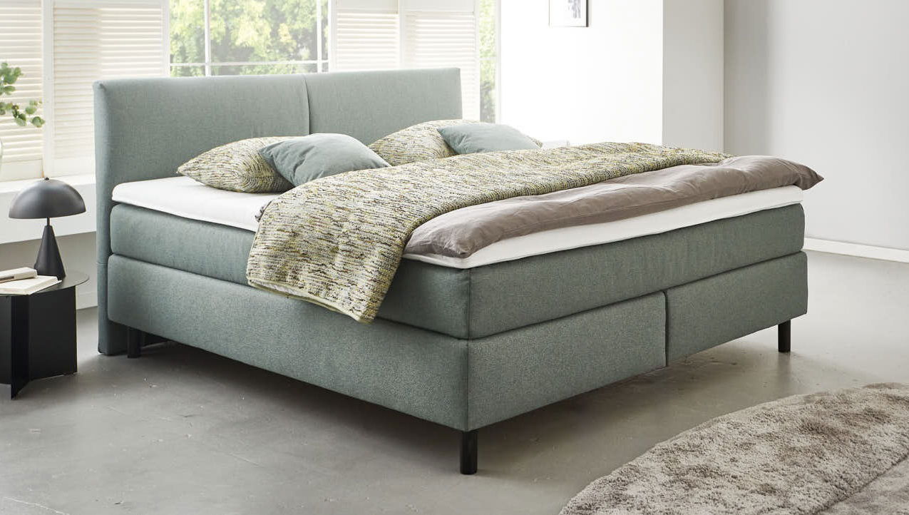 Boxspringbett BX 180/200 in modernem Design, seitliche Perspektive, mit grauem Stoffbezug und gemütlicher Bettwäsche.