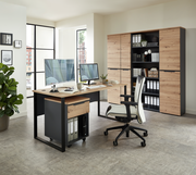Moderner Schreibtisch mit Holz- und Metallelementen, ausgestattet mit zwei Monitoren, einem Rollcontainer und einem ergonomischen Bürostuhl, in einem hellen Büro mit Blick auf Aktenschränke im Hintergrund.