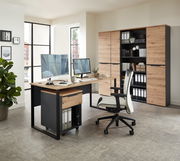 Moderner Schreibtisch mit Holz- und Metallelementen, ausgestattet mit zwei Monitoren, einem Rollcontainer und einem ergonomischen Bürostuhl, in einem hellen Büro mit Blick auf Aktenschränke im Hintergrund.