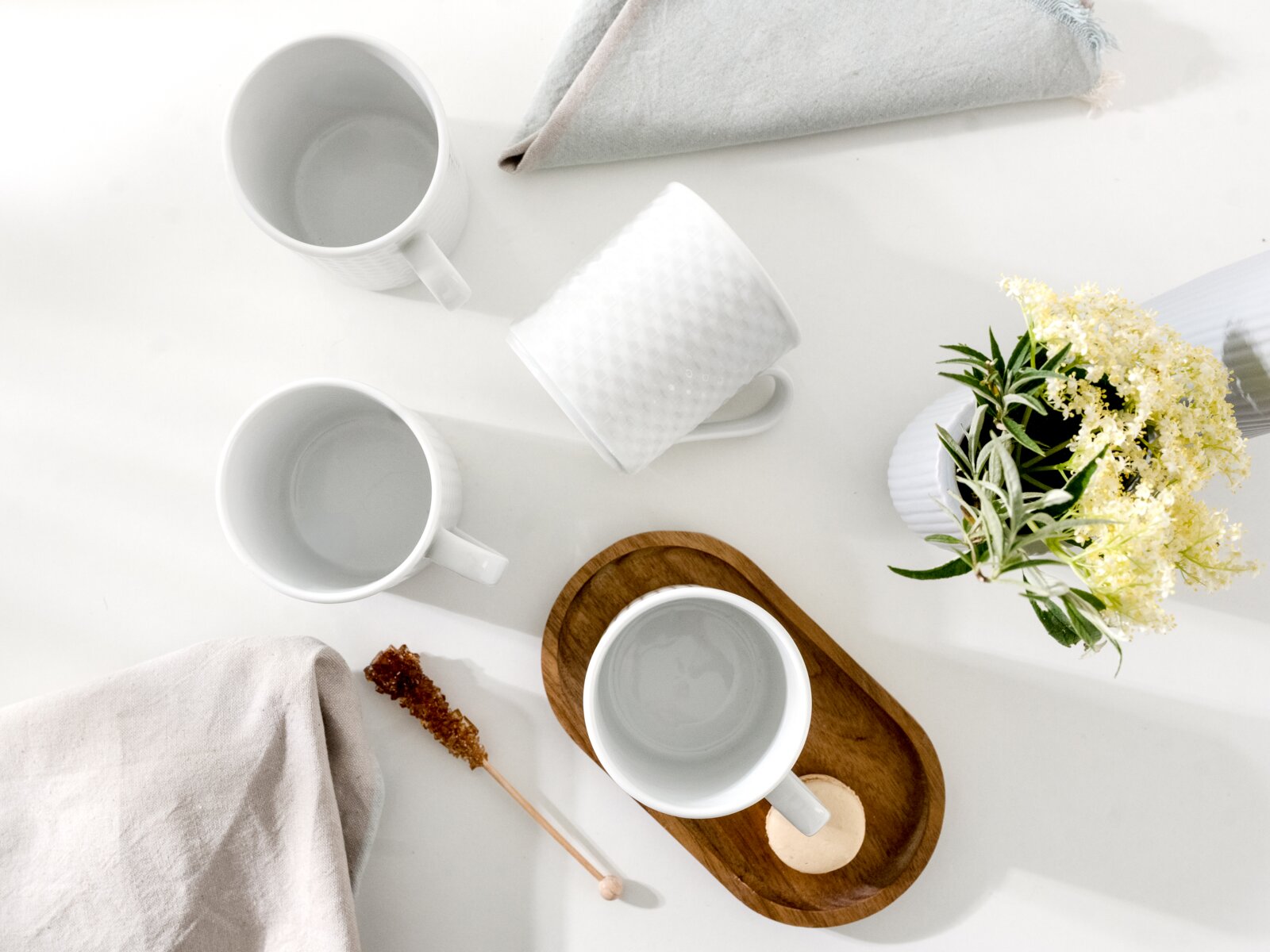 Drei weiße Keramik-Kaffeetassen und ein Holztablett mit Tasse auf weißem Tisch, daneben Stoffservietten, Kandiszucker und eine Vase mit hellen Blumen, fotografiert aus der Vogelperspektive (Top-Down).