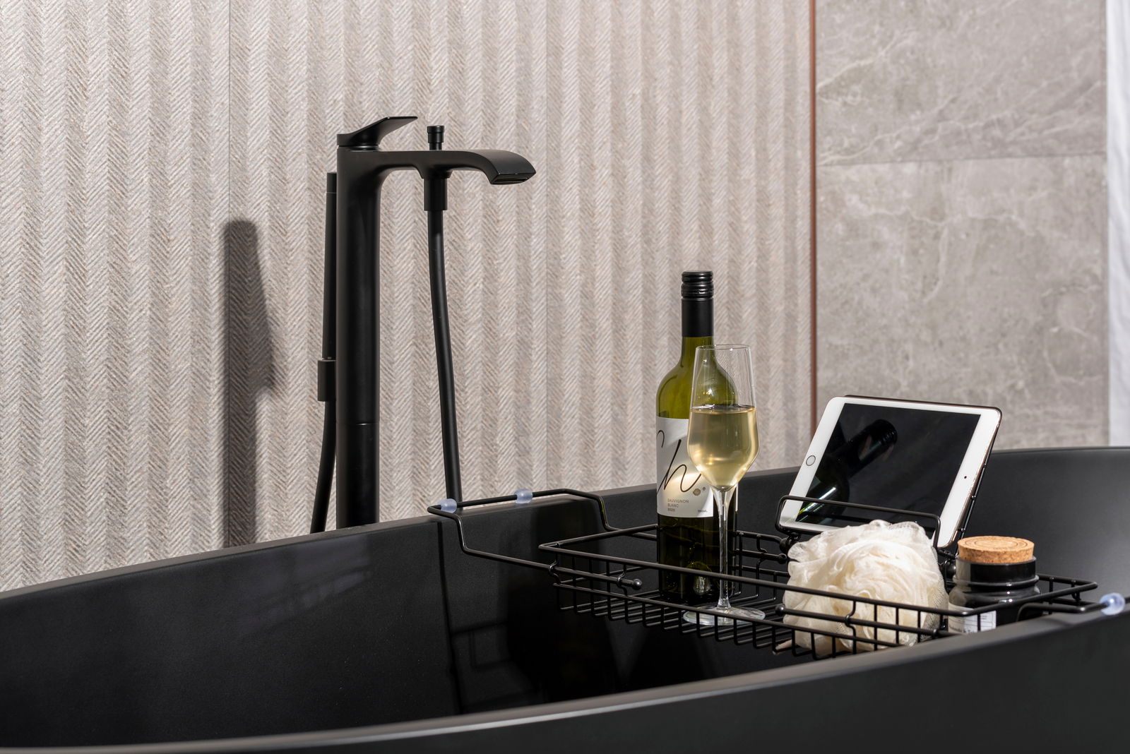 Badablage über einer schwarzen Badewanne mit einer Flasche Wein, einem Weinglas, einem Tablet und einem Duschschwamm, seitliche Perspektive.