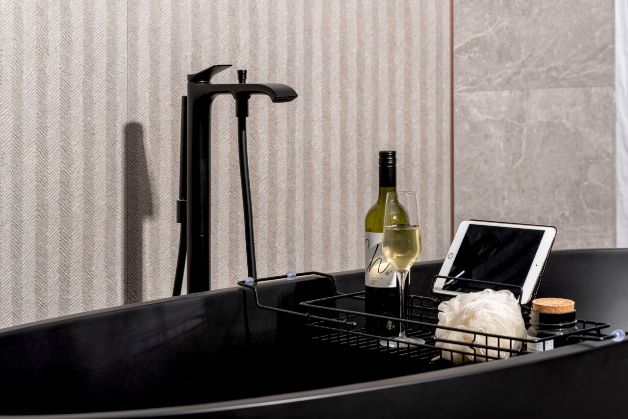Badablage über einer schwarzen Badewanne mit einer Flasche Wein, einem Weinglas, einem Tablet und einem Duschschwamm, seitliche Perspektive.