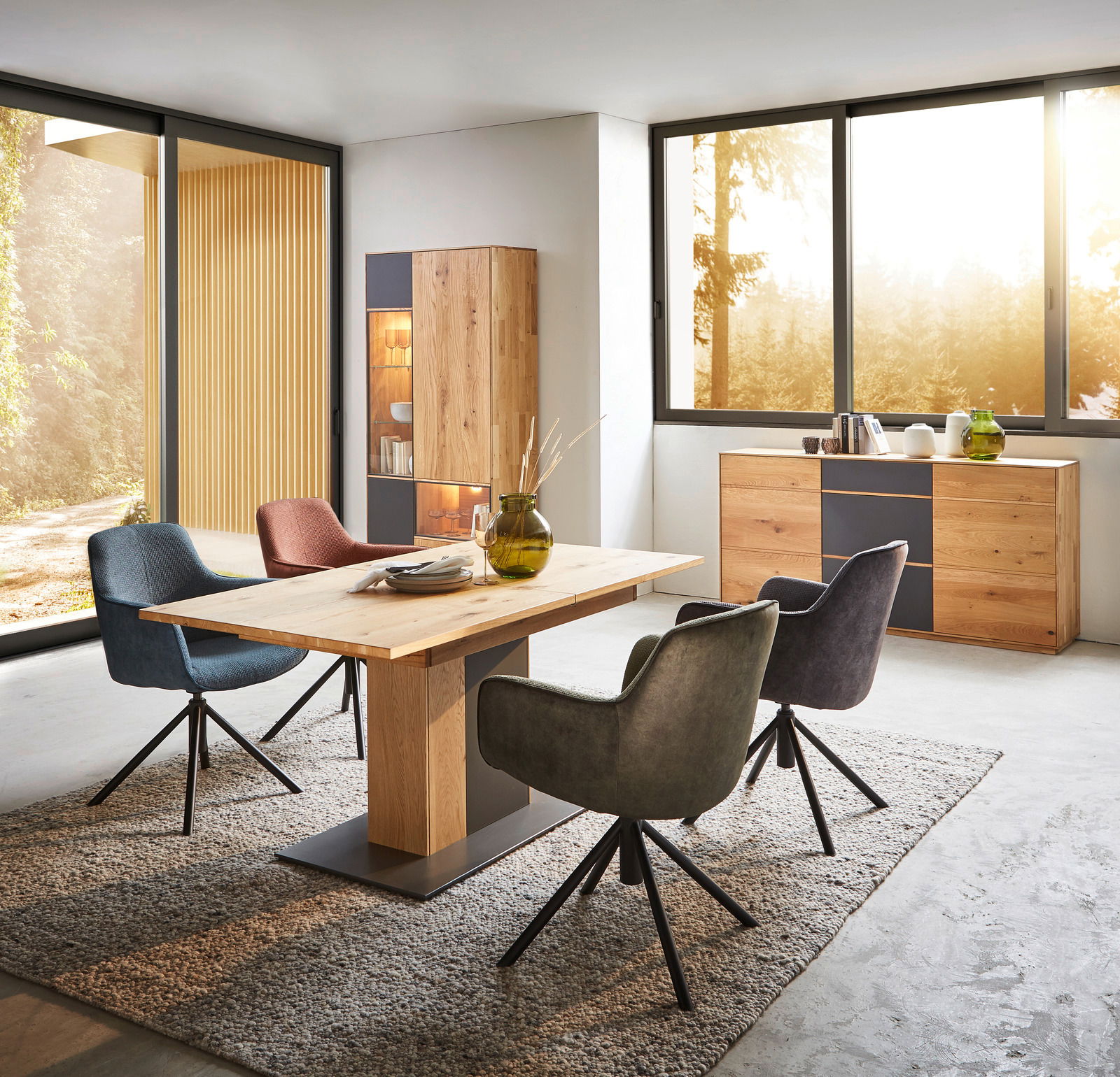 Waldwelt Sideboard 2-türig AVANTI Modernes Esszimmer mit einem hölzernen Sideboard, zwei Türen, in einer hellen, von großen Fenstern umgebenen Umgebung, mit einem Esstisch und bunten Stühlen, seitliche Perspektive.