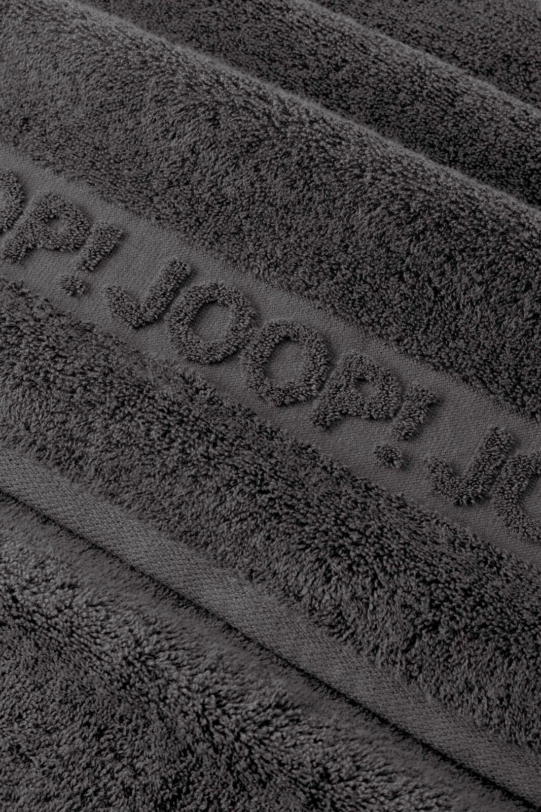 JOOP! Frottierset 5-teilig STUDIO Nahaufnahme eines dunkelgrauen Frottee-Handtuchs mit eingewebtem Schriftzug „TOM TAILOR“, schräg von oben fotografiert.