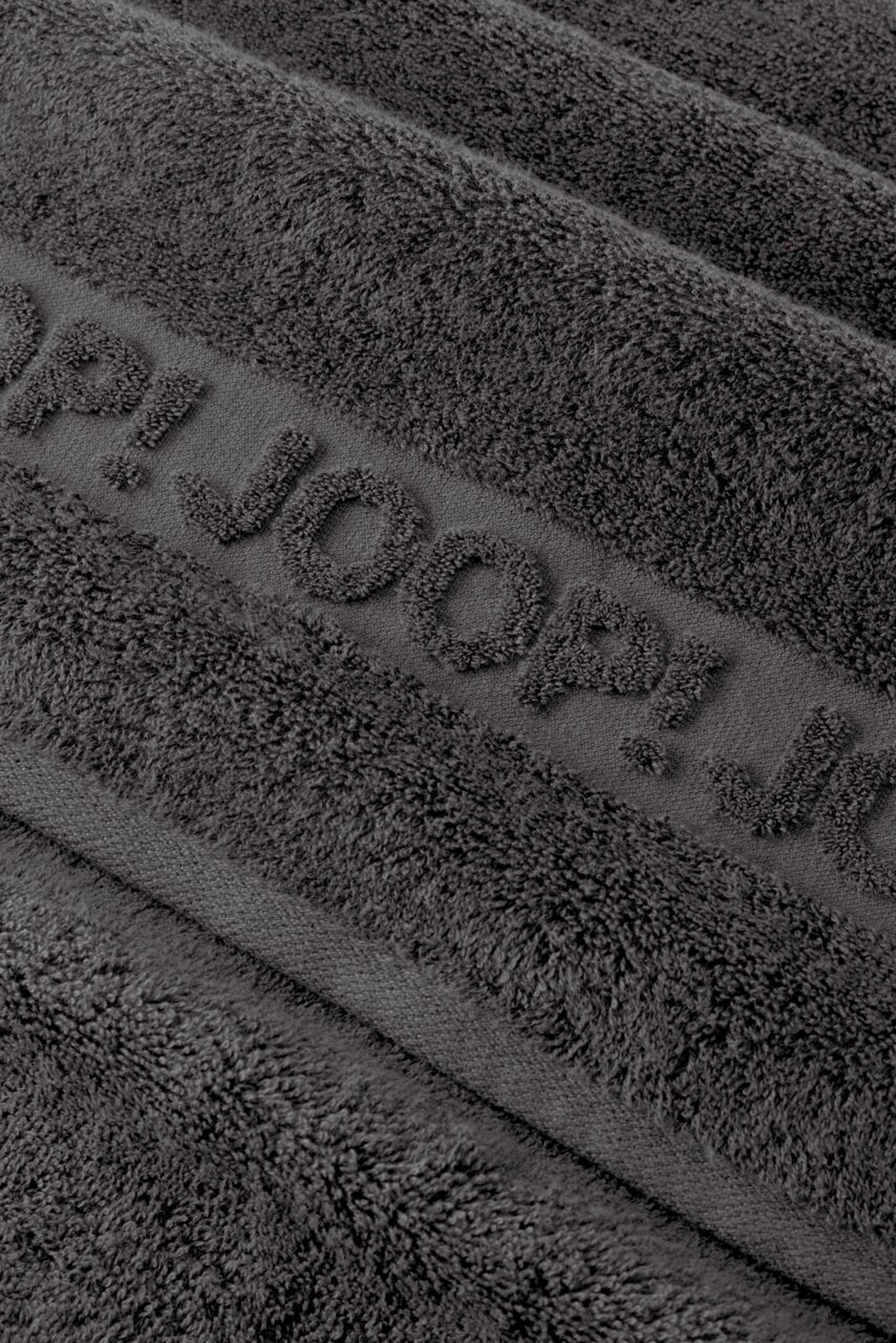 JOOP! Frottierset 5-teilig STUDIO Nahaufnahme eines dunkelgrauen Frottee-Handtuchs mit eingewebtem Schriftzug „TOM TAILOR“, schräg von oben fotografiert.