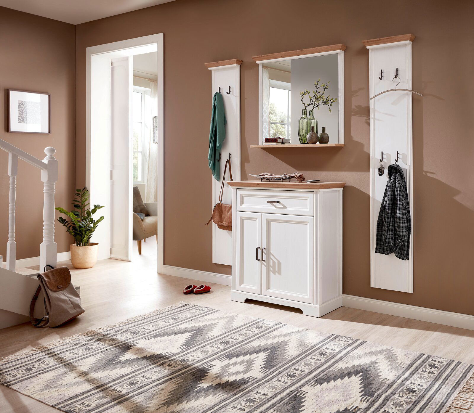 Fluransicht mit weißem Garderoben-Set, bestehend aus zwei schmalen Garderobenpaneelen, einem Spiegel und einem Schuhschrank, auf hellem Holzboden vor brauner Wand.