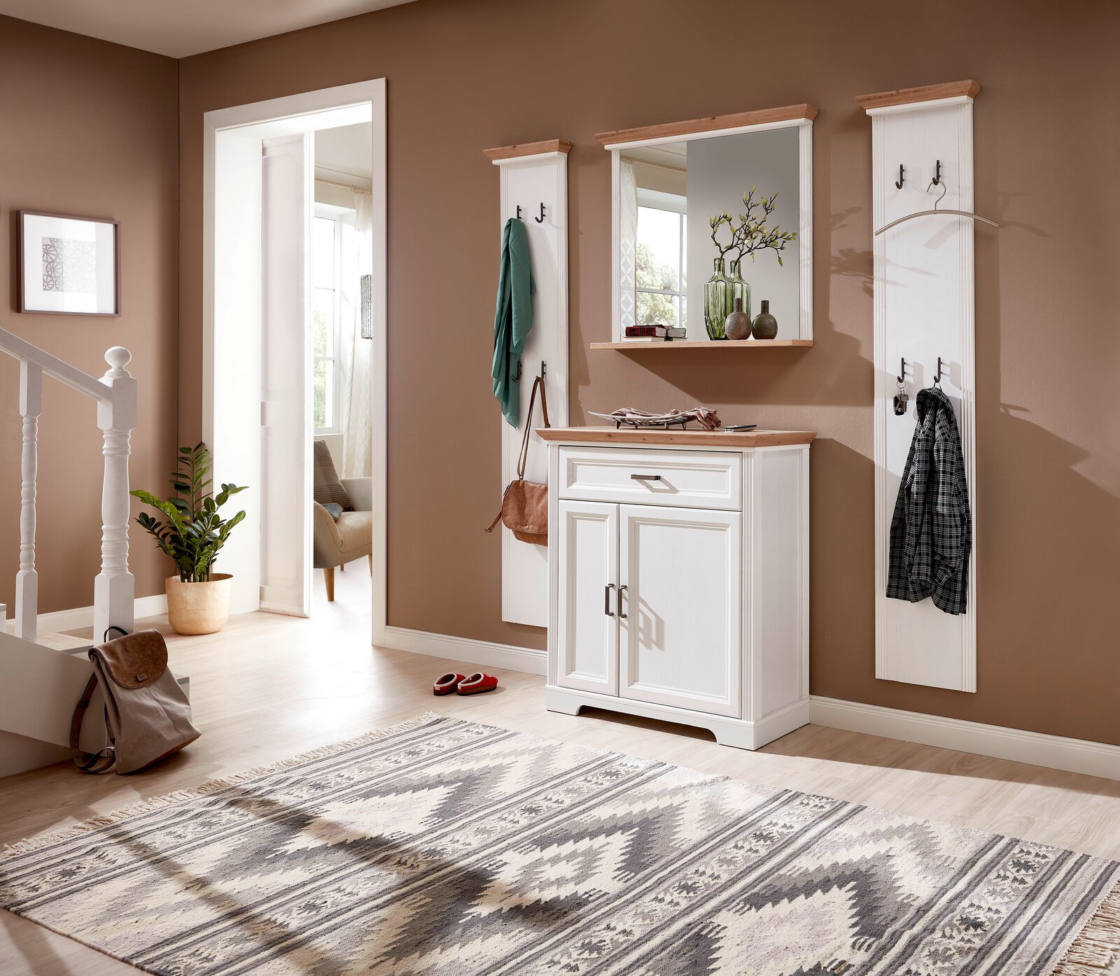 Fluransicht mit weißem Garderoben-Set, bestehend aus zwei schmalen Garderobenpaneelen, einem Spiegel und einem Schuhschrank, auf hellem Holzboden vor brauner Wand.