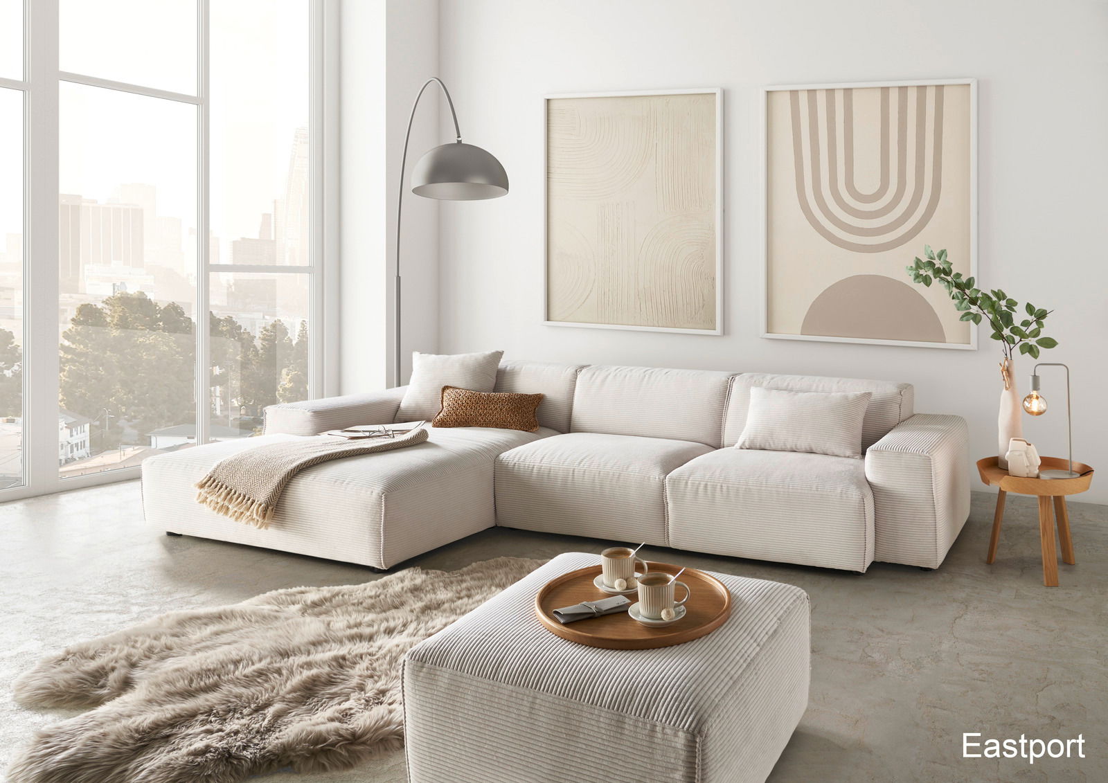 Lounge Handmade Ecksofa LH-EDINA Moderne Wohnzimmeransicht mit einer großen, hellen Rundecke, dekoriert mit Kissen und Decken, einem flauschigen Teppich und einem Beistelltisch mit Lampe. Große Fenster bieten einen Blick auf die Stadt.