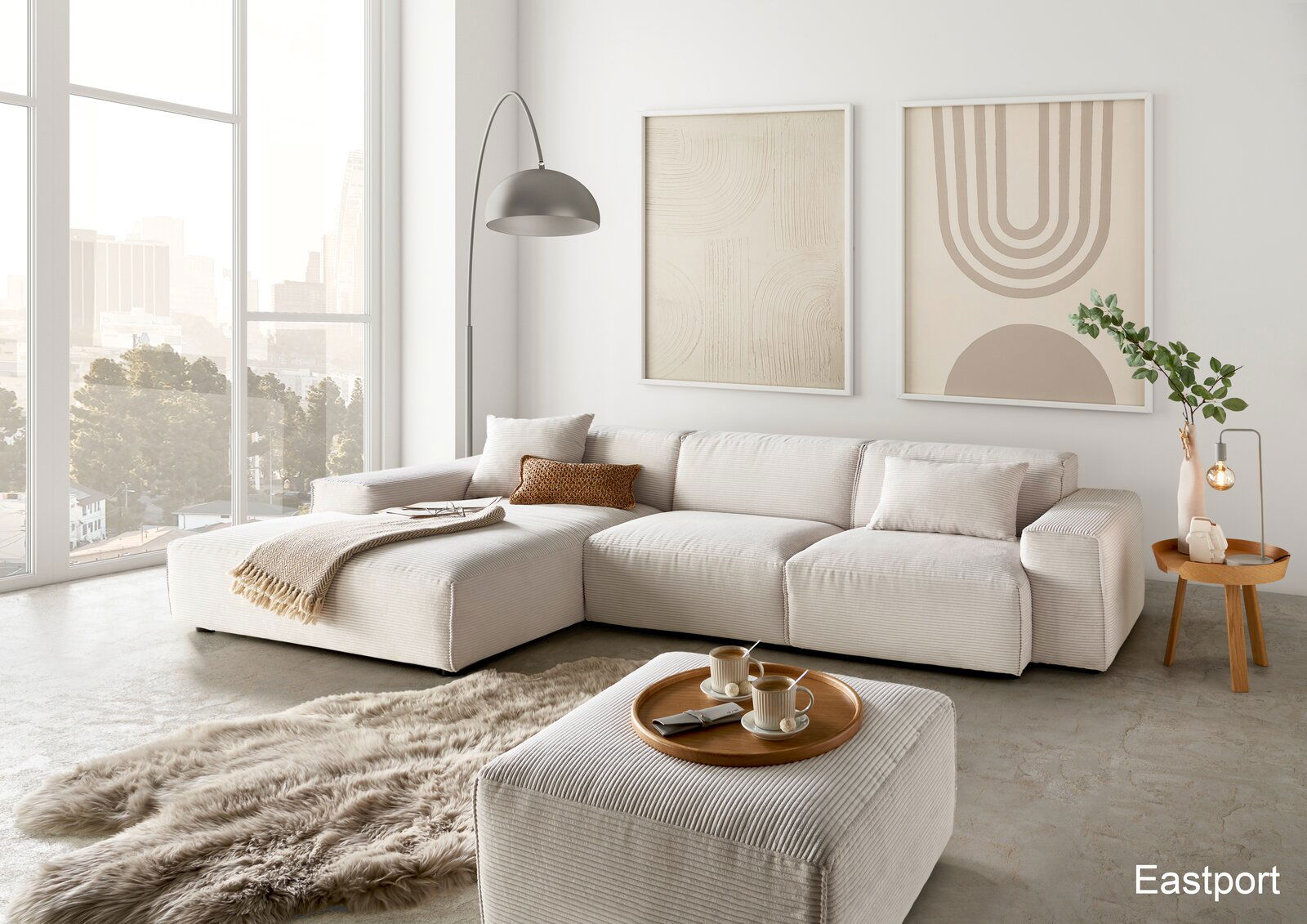 Moderne Wohnzimmeransicht mit einer großen, hellen Rundecke, dekoriert mit Kissen und Decken, einem flauschigen Teppich und einem Beistelltisch mit Lampe. Große Fenster bieten einen Blick auf die Stadt.