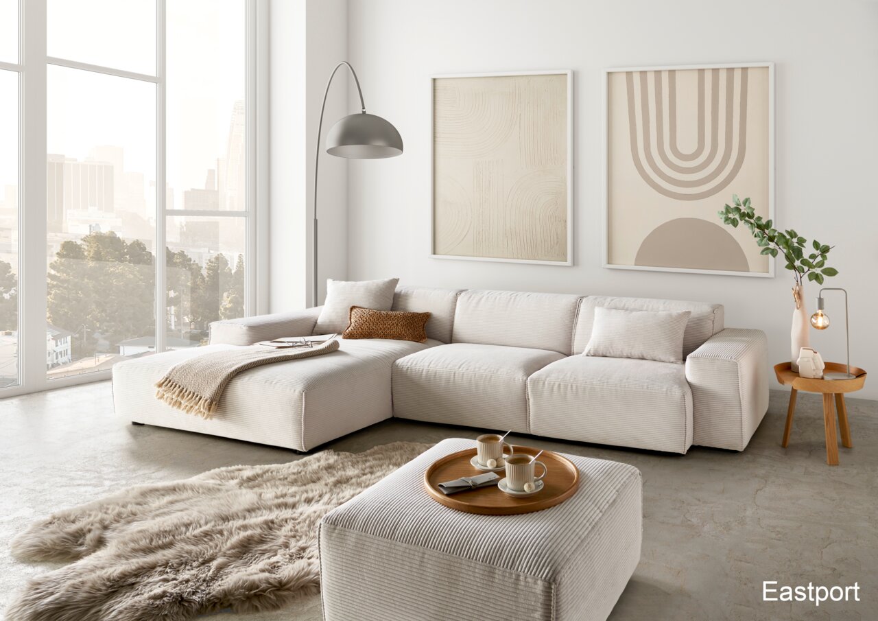 Lounge Handmade Ecksofa LH-EDINA Moderne Wohnzimmeransicht mit einer großen, hellen Rundecke, dekoriert mit Kissen und Decken, einem flauschigen Teppich und einem Beistelltisch mit Lampe. Große Fenster bieten einen Blick auf die Stadt.