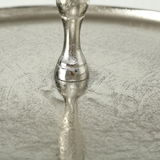 Nahaufnahme einer silbernen, strukturierten Oberfläche mit einem dekorativen Ständer in der Mitte, von oben betrachtet.