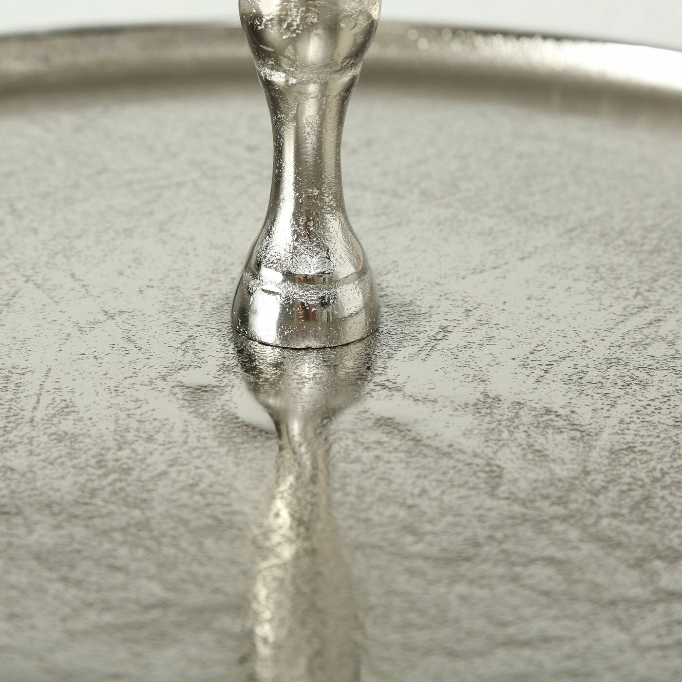 Nahaufnahme einer silbernen, strukturierten Oberfläche mit einem dekorativen Ständer in der Mitte, von oben betrachtet.