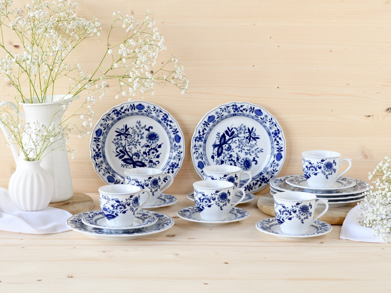 Blau-weißes Porzellan-Kaffeeservice mit floralem Muster, Tassen auf Untertassen und Teller-Set vor hellen Holzhintergrund, dekoriert mit weißen Blumenvasen, Frontansicht auf Tischhöhe