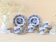 Blau-weißes Porzellan-Kaffeeservice mit floralem Muster, Tassen auf Untertassen und Teller-Set vor hellen Holzhintergrund, dekoriert mit weißen Blumenvasen, Frontansicht auf Tischhöhe