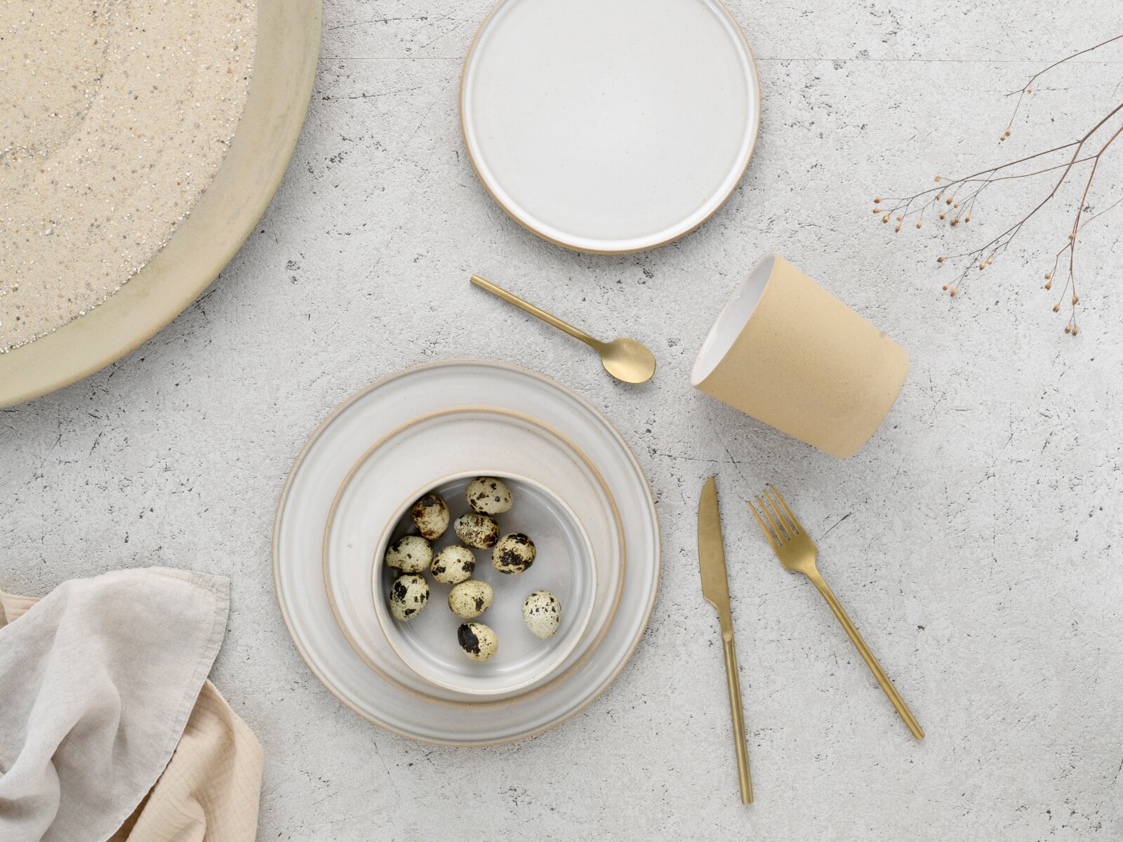Minimalistisches Geschirr-Set in Beige und Weiß mit Teller, Schale mit Wachteleiern, Becher und goldfarbenem Besteck auf heller Steinoberfläche, Draufsicht von oben (Flatlay).