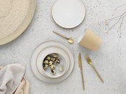 Minimalistisches Geschirr-Set in Beige und Weiß mit Teller, Schale mit Wachteleiern, Becher und goldfarbenem Besteck auf heller Steinoberfläche, Draufsicht von oben (Flatlay).