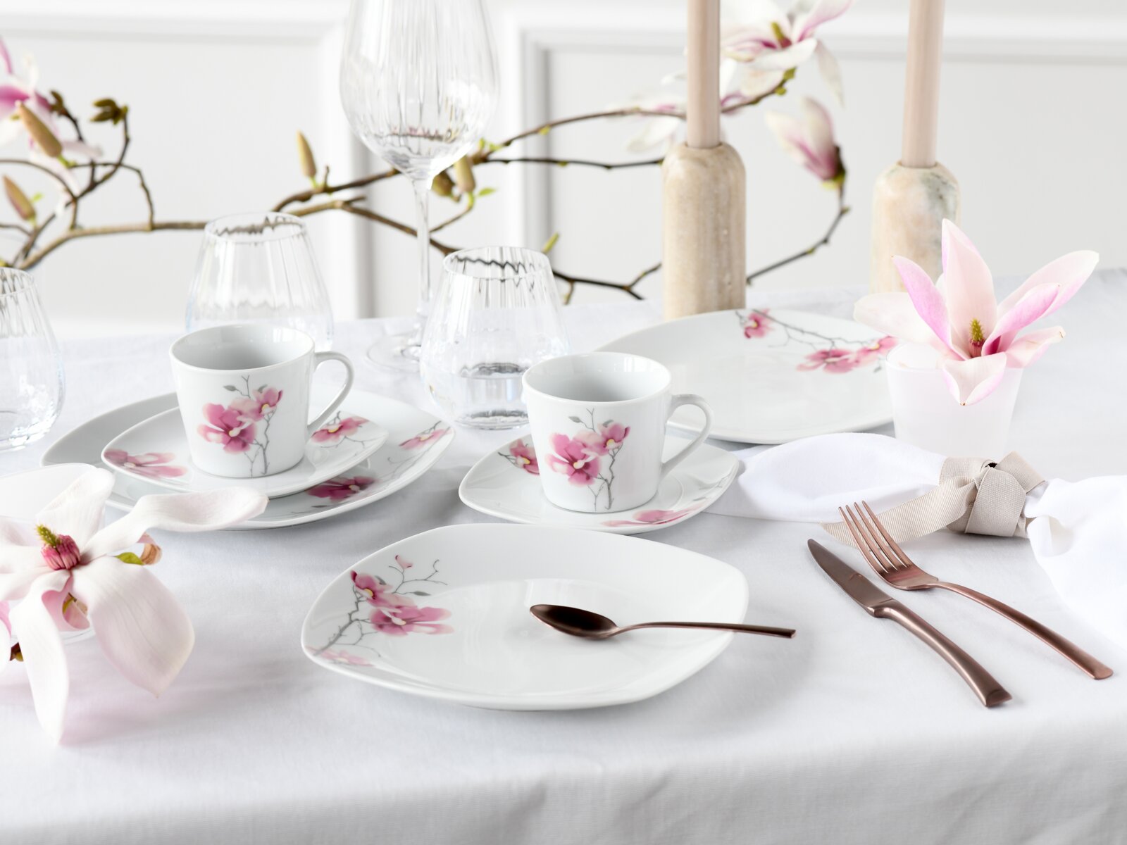 Elegant gedeckter Tisch mit weißem Porzellan-Geschirrset mit rosa Blütenmotiv (Tassen, Teller), Kristallgläsern und roségoldenem Besteck; Ansicht schräg von vorne auf Augenhöhe.
