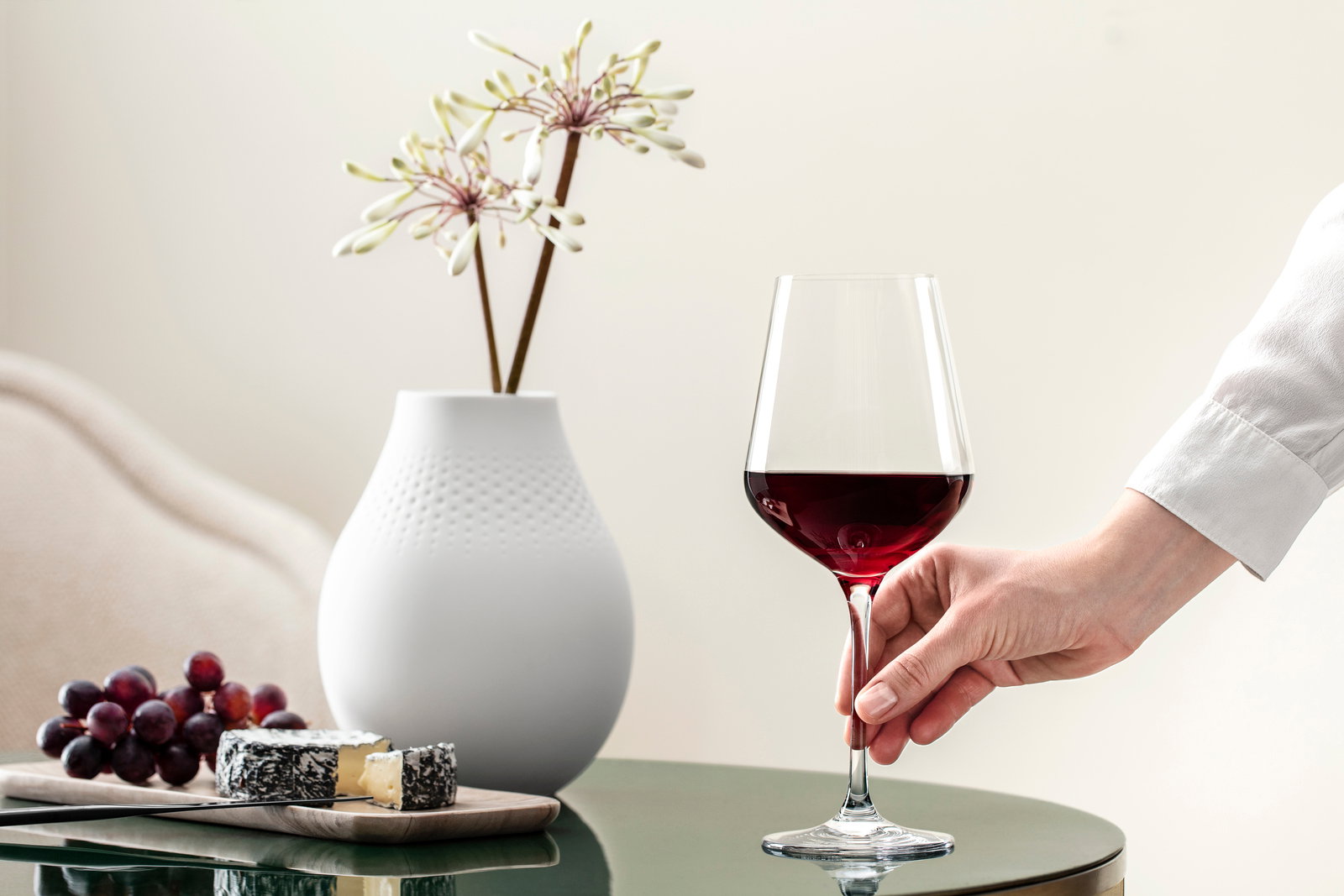 Villeroy & Boch Set van 4 Bordeauxglazen LA DAVINA Ein elegantes Bordeauxkelch-Glas, gefüllt mit Rotwein, wird von einer Hand gehalten. Im Hintergrund sind eine weiße Vase mit Blumen und ein Käsebrett mit Trauben zu sehen. Perspektive: seitlich.