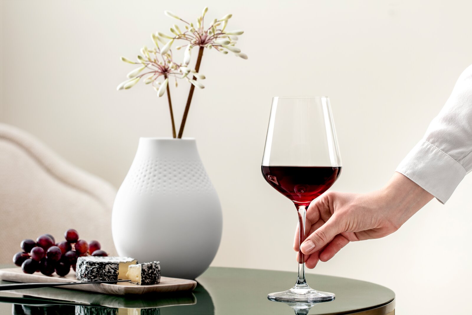 Villeroy & Boch Set van 4 Bordeauxglazen LA DAVINA Een elegant bordeauxglas, gevuld met rode wijn, wordt door een hand vastgehouden. Op de achtergrond zijn een witte vaas met bloemen en een kaasplankje met druiven te zien. Perspectief: zijaanzicht.