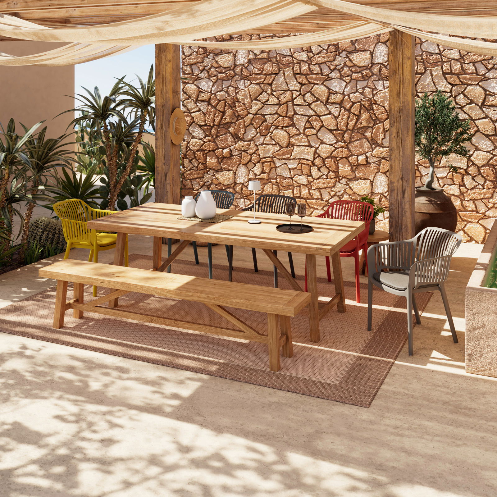 Gartentisch aus Holz mit passender Bank und bunten Stühlen, umgeben von Pflanzen, in einer sonnigen Terrasse mit Steinwand im Hintergrund, aus seitlicher Perspektive.