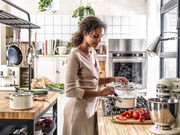 Frau kocht in moderner Küche mit dampfendem Topfset in almond white auf dem Herd, seitliche Perspektive.