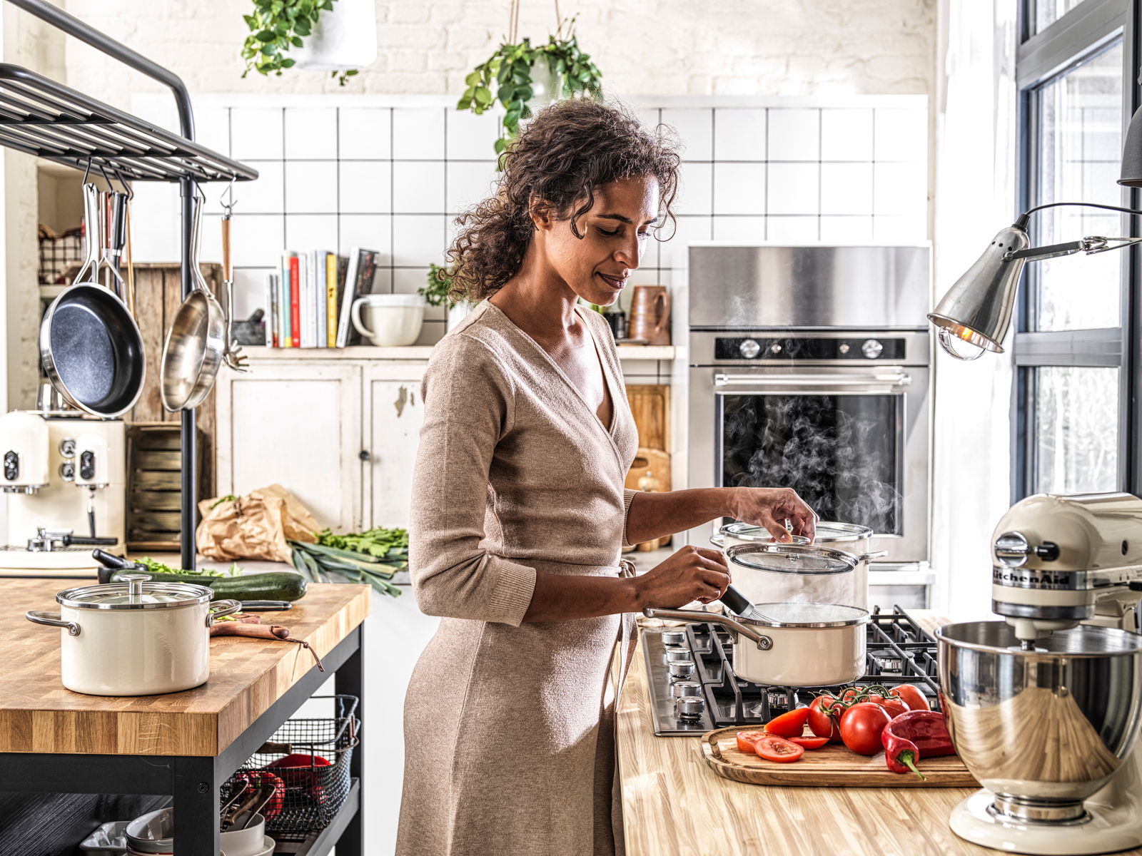 KitchenAid Topfset 4-tlg. COOKWARE Frau kocht in moderner Küche mit dampfendem Topfset in almond white auf dem Herd, seitliche Perspektive.