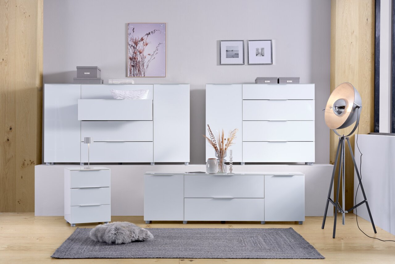 Moderne weiße Möbelkollektion in einem stilvollen Raum, bestehend aus einem TV-Lowboard, einer Nachtkonsole und mehreren Kommoden, aus der Frontalperspektive betrachtet.