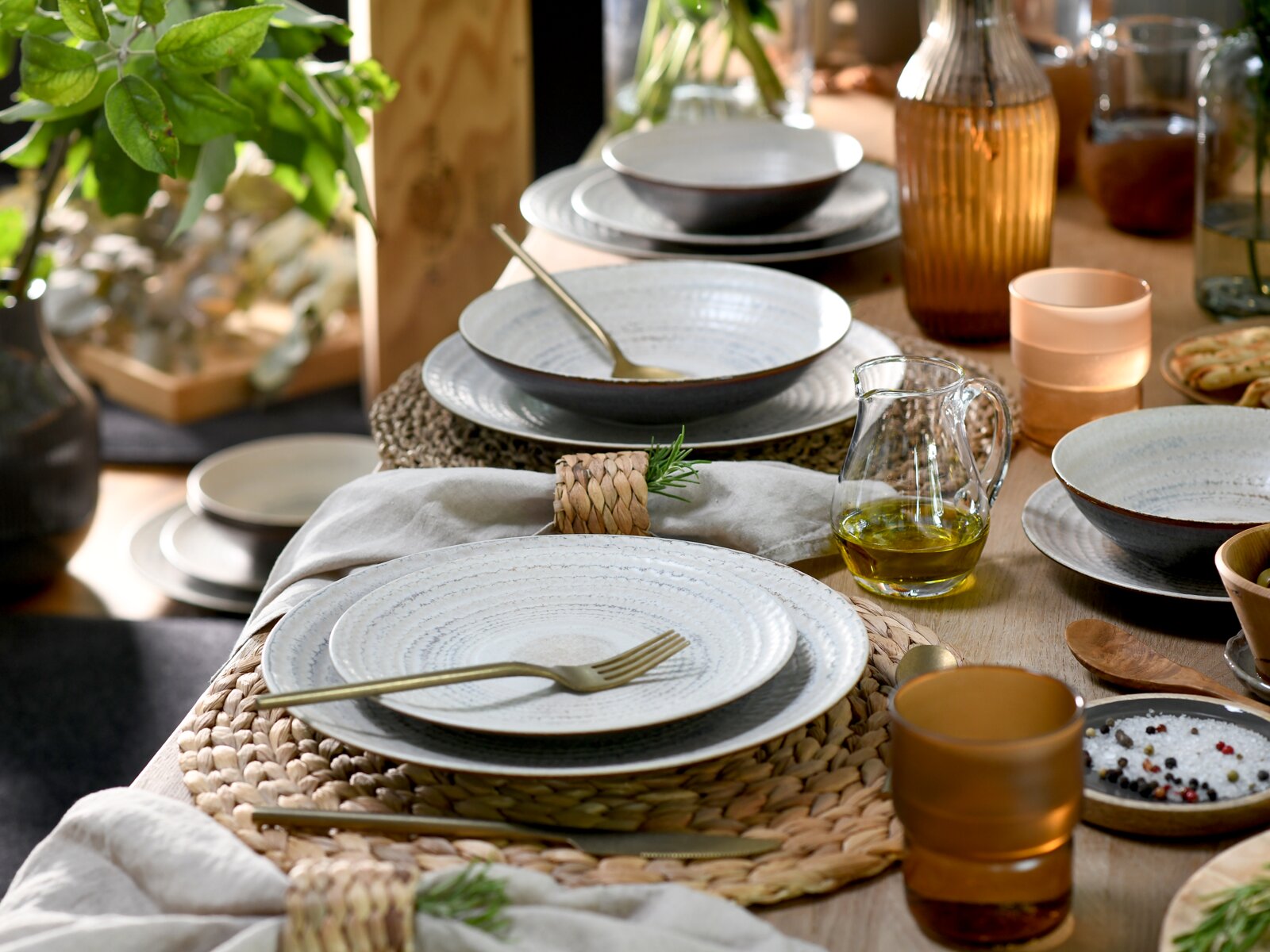 Schlicht gedeckter Holztisch mit graublauem Keramikgeschirr (Teller und Schalen), goldenem Besteck, geflochtenen Platzsets, Leinenservietten, Glasflaschen und Kerzenlicht, fotografiert aus leicht erhöhter schräger Perspektive von oben.